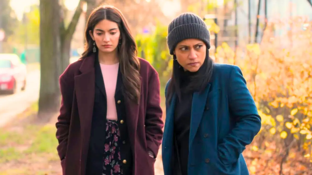Two Indian women walking together in an autumn park with serious expressions, one in maroon coat, other in teal coat and beanie