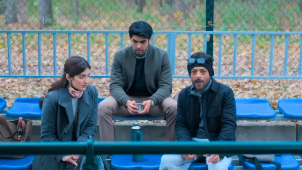 Three Indian people in serious discussion on park bleachers: woman in coat, man in green jacket, bearded man in beanie, autumn outdoor setting